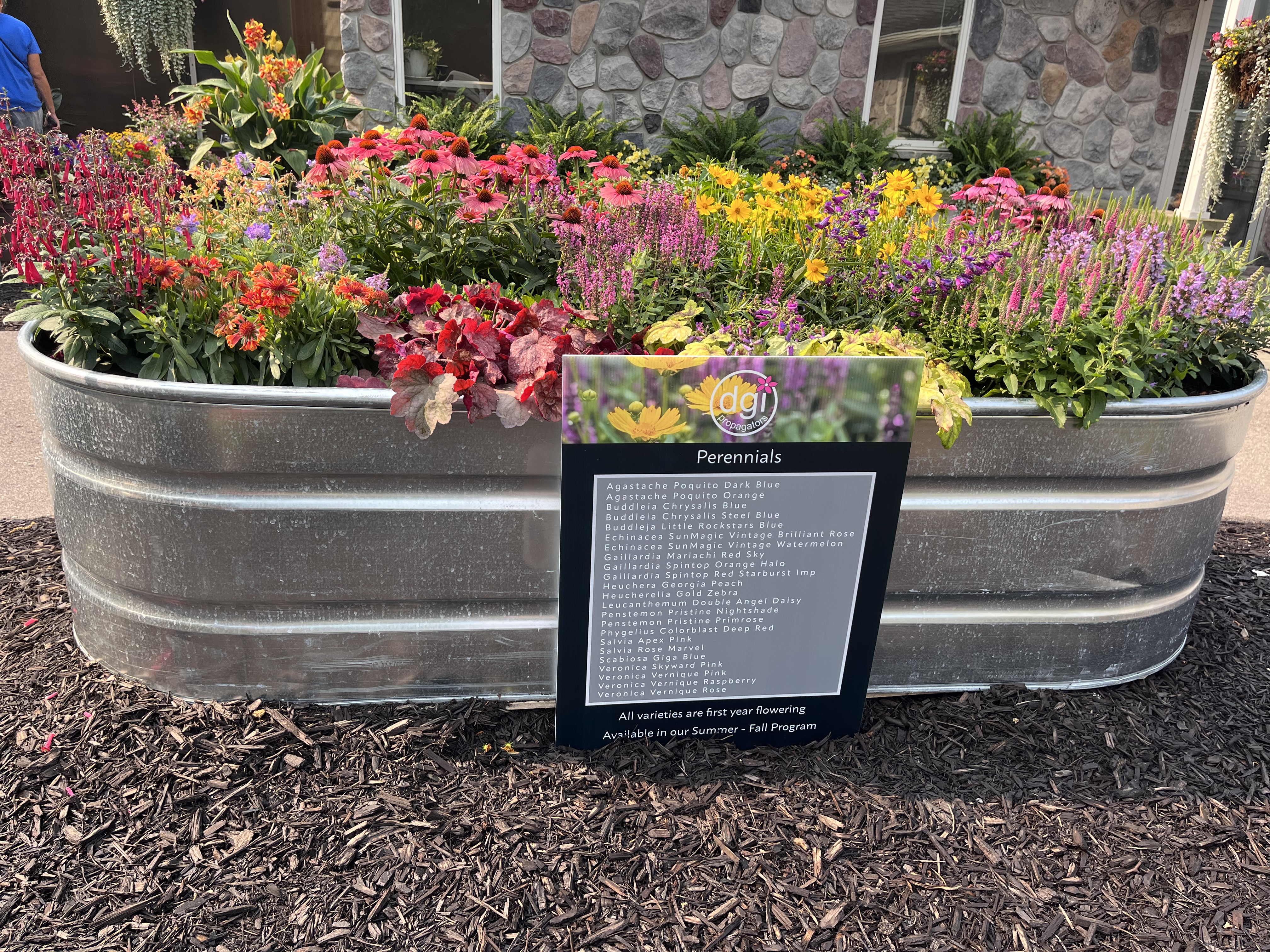A large galvanized metal planter filled with a colorful mix of perennial flowers, including pink, purple, orange, yellow, and red blooms. In front, a sign lists the perennial varieties featured.